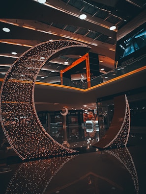 A crescent-shaped installation adorned with numerous small lights stands in a modern indoor space. The ambient lighting creates a warm and inviting atmosphere. The ceiling features contemporary design elements with recessed lighting and round fixtures. Reflections on the polished floor enhance the aesthetic appeal of the setting.