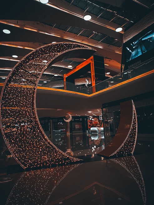 A crescent-shaped installation adorned with numerous small lights stands in a modern indoor space. The ambient lighting creates a warm and inviting atmosphere. The ceiling features contemporary design elements with recessed lighting and round fixtures. Reflections on the polished floor enhance the aesthetic appeal of the setting.