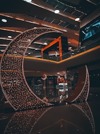 A crescent-shaped installation adorned with numerous small lights stands in a modern indoor space. The ambient lighting creates a warm and inviting atmosphere. The ceiling features contemporary design elements with recessed lighting and round fixtures. Reflections on the polished floor enhance the aesthetic appeal of the setting.