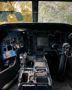 A dilapidated aircraft cockpit with missing and damaged control panels. The windshield is cracked, allowing view of overgrown vegetation outside. Dust and debris cover the instrument consoles, giving an impression of abandonment and decay.