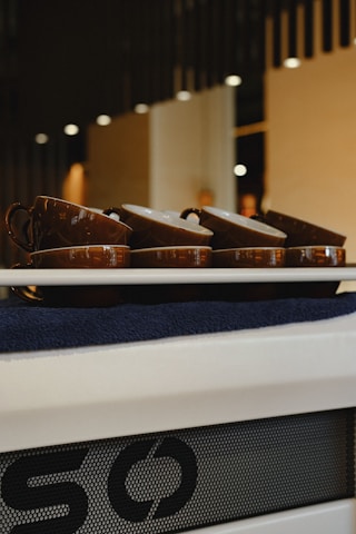A set of brown cups is neatly arranged on a tray, placed against a modern, softly lit interior backdrop. The focus is on the cups, which seem to be on a towel, with a portion of a logo or design visible beneath the tray.
