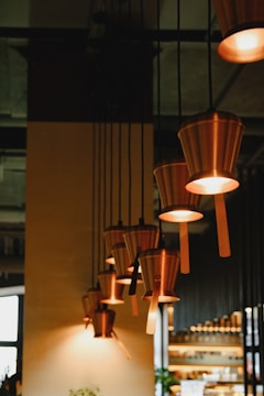 Several pendant lights with warm, soft glows hang in a row, resembling upside-down metal buckets. The setting appears to be a stylish and modern interior space, possibly a restaurant or cafe, with dark and warm tones complementing the cozy atmosphere.