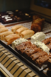 An assortment of pastries arranged on a display tray. The foreground features various types of pastries with different toppings and glazes, including chocolate and white chocolate items with nuts. Behind them, more baked goods are arranged in rows, showcasing a variety of textures and colors.