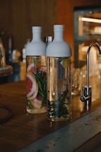 Two glass bottles filled with water are placed on a wooden counter. One bottle contains slices of grapefruit and sprigs of rosemary, while the other contains lemon slices and leafy herbs. The setting appears to be a kitchen or bar area with a metallic faucet in the background.