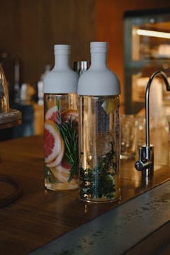 Two glass bottles filled with water are placed on a wooden counter. One bottle contains slices of grapefruit and sprigs of rosemary, while the other contains lemon slices and leafy herbs. The setting appears to be a kitchen or bar area with a metallic faucet in the background.