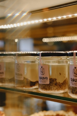 Several glass jars filled with layered dessert are arranged on a glass shelf. The jars have a label with the word 'takava' and a logo. The dessert appears layered with creamy and crumbly textures.