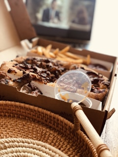 A close-up of a pizza in a takeout box, with some fries on the side and a plastic lid with sauce resting on top. There is a wicker basket in the foreground, and a blurred television screen can be seen in the background, indicating a cozy meal setting.