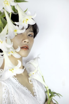 A person with short dark hair is partially obscured by white lilies, wearing a delicate white lace garment. The background is a soft, neutral color, and the overall composition presents an aesthetic blend of floral elements and human expression.