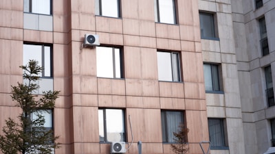 A modern apartment building facade with a uniform pattern of glass windows and beige panels. Air conditioning units are mounted on some of the windows, and pine trees grow in front of the building.