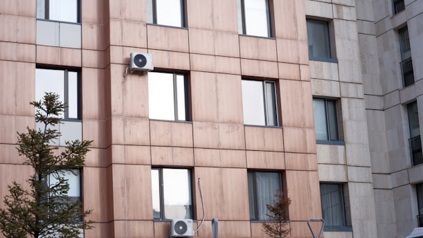A modern apartment building facade with a uniform pattern of glass windows and beige panels. Air conditioning units are mounted on some of the windows, and pine trees grow in front of the building.