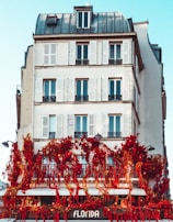 A multi-story building with classic architecture features white walls and multiple windows with shutters. The lower level of the building is adorned with vibrant red flowers and greenery, creating a striking contrast against the white facade. The sign 'Florida' is visible at the bottom, suggesting a shop or café.