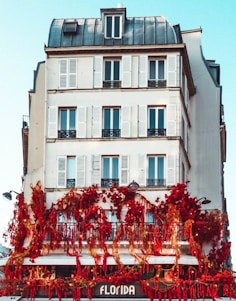 A multi-story building with classic architecture features white walls and multiple windows with shutters. The lower level of the building is adorned with vibrant red flowers and greenery, creating a striking contrast against the white facade. The sign 'Florida' is visible at the bottom, suggesting a shop or café.