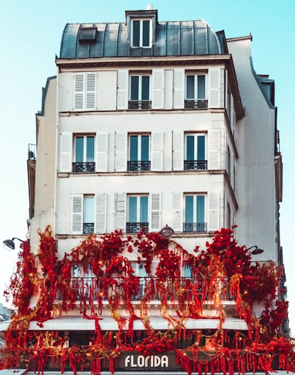 A multi-story building with classic architecture features white walls and multiple windows with shutters. The lower level of the building is adorned with vibrant red flowers and greenery, creating a striking contrast against the white facade. The sign 'Florida' is visible at the bottom, suggesting a shop or café.