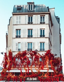 A multi-story building with classic architecture features white walls and multiple windows with shutters. The lower level of the building is adorned with vibrant red flowers and greenery, creating a striking contrast against the white facade. The sign 'Florida' is visible at the bottom, suggesting a shop or café.