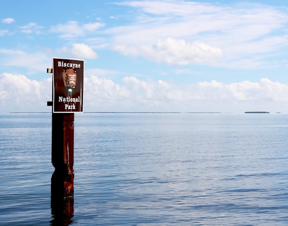 A signpost stands in calm waters, displaying the name 'Biscayne National Park' against a backdrop of a vast expanse of water and a partly cloudy sky. The horizon stretches across the image with small distant landmasses.