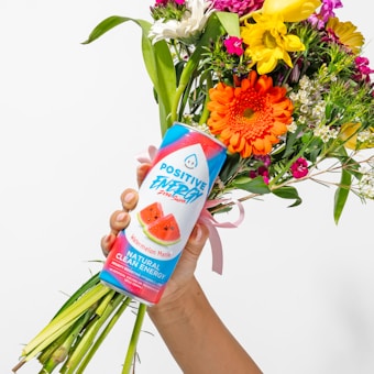 A hand holds a vibrant bouquet of various colorful flowers along with a can of energy drink labeled 'Positive Energy'. The flowers include orange, yellow, pink, and white blooms, tied together with a pink ribbon. The can features bright colors and images of watermelon slices.