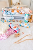 A neatly packed suitcase is open on a bed, containing various items such as clothing and orange-colored spray cans. Several canned beverages with vibrant labels are scattered across the white bedspread alongside a small bottle of alcohol. A gold chain necklace lies near a pink floral-patterned fabric.