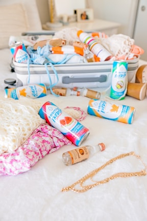 A neatly packed suitcase is open on a bed, containing various items such as clothing and orange-colored spray cans. Several canned beverages with vibrant labels are scattered across the white bedspread alongside a small bottle of alcohol. A gold chain necklace lies near a pink floral-patterned fabric.