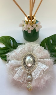 A green vase with golden accents is adorned with white flowers and reed sticks. In front, a decorative object featuring white lace and pearls with a central pearl-like stone is complemented by two large leaves.