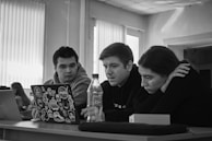 Three people are seated at a table, focused on a laptop covered with various stickers including one from NASA. They appear to be collaborating or studying together. A bottle is placed on the table, and there is a box and a closed laptop nearby. The room is well-lit with natural light coming through vertical blinds.