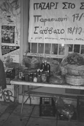 A monochrome image depicts a market stall with a table displaying jars and bottles, possibly homemade preserves and pickles. Above the stall, a banner in Greek advertises a market event. Posters are visible on the wall in the background.