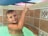 A young child with short hair, wet from swimming, is in a pool. The child is pointing upward with one finger and smiling slightly. The pool has decorative tile walls with geometric patterns, and the water appears clear.