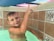 A young child with short hair, wet from swimming, is in a pool. The child is pointing upward with one finger and smiling slightly. The pool has decorative tile walls with geometric patterns, and the water appears clear.