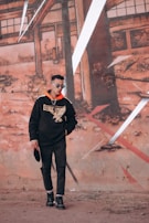 A man wearing a black hoodie with a large graphic design stands in front of an urban mural. He has sunglasses and is holding a hat, posing casually against a background of abstract, geometric wall art with earthy tones.