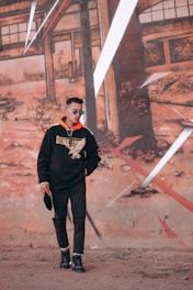 A man wearing a black hoodie with a large graphic design stands in front of an urban mural. He has sunglasses and is holding a hat, posing casually against a background of abstract, geometric wall art with earthy tones.