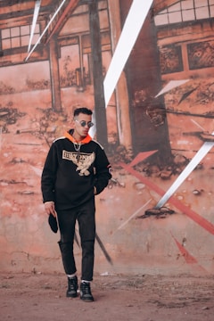 A man wearing a black hoodie with a large graphic design stands in front of an urban mural. He has sunglasses and is holding a hat, posing casually against a background of abstract, geometric wall art with earthy tones.