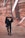 A man wearing a black hoodie with a large graphic design stands in front of an urban mural. He has sunglasses and is holding a hat, posing casually against a background of abstract, geometric wall art with earthy tones.
