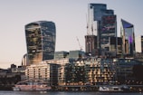 A modern cityscape featuring several high-rise buildings with unique architectural designs. The structures include the Walkie Talkie skyscraper, with its distinctive curved shape and glass facade. Cranes are visible, indicating ongoing construction. In the foreground, a river with moored boats and lit-up buildings reflect the evening light.