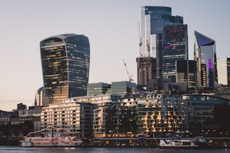 A modern cityscape featuring several high-rise buildings with unique architectural designs. The structures include the Walkie Talkie skyscraper, with its distinctive curved shape and glass facade. Cranes are visible, indicating ongoing construction. In the foreground, a river with moored boats and lit-up buildings reflect the evening light.