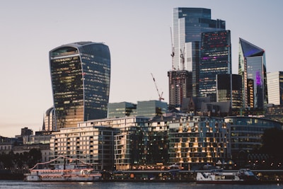 A modern cityscape featuring several high-rise buildings with unique architectural designs. The structures include the Walkie Talkie skyscraper, with its distinctive curved shape and glass facade. Cranes are visible, indicating ongoing construction. In the foreground, a river with moored boats and lit-up buildings reflect the evening light.