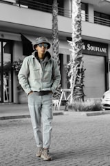 A person wearing casual clothing, including a denim jacket and jeans, along with a bucket hat and sunglasses, walks along a cobblestone street. The background features palm trees and a building with signage.