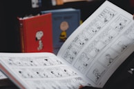 A person is holding an open comic strip book featuring black and white illustrations. In the background, there are two hardcover books with cartoon covers standing upright, one red with an image of a dog and another with a yellow character.