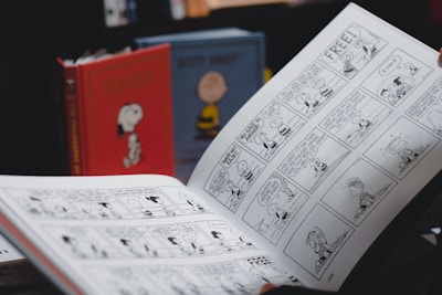 A person is holding an open comic strip book featuring black and white illustrations. In the background, there are two hardcover books with cartoon covers standing upright, one red with an image of a dog and another with a yellow character.