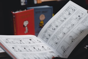 A person is holding an open comic strip book featuring black and white illustrations. In the background, there are two hardcover books with cartoon covers standing upright, one red with an image of a dog and another with a yellow character.