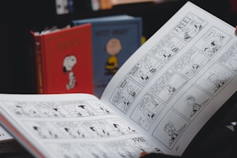 A person is holding an open comic strip book featuring black and white illustrations. In the background, there are two hardcover books with cartoon covers standing upright, one red with an image of a dog and another with a yellow character.