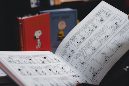 A person is holding an open comic strip book featuring black and white illustrations. In the background, there are two hardcover books with cartoon covers standing upright, one red with an image of a dog and another with a yellow character.