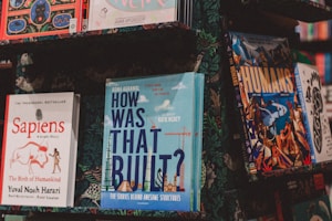 A collection of books displayed on a shelf, featuring a variety of titles and colorful covers. Prominent books include 'Sapiens' with a red and white design, 'How Was That Built?' with a blue cover and construction elements, and 'The Humans' with an illustrated scene and bold typography. The surrounding books and background have a muted floral pattern.