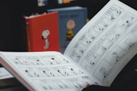 An open comic book displaying black and white comic strips. In the background, there are two hardcover books with colorful covers featuring characters from the comic. The image suggests a setting focused on reading or comic collection.