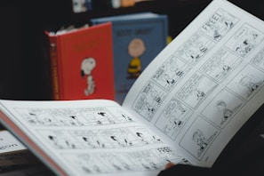 An open comic book displaying black and white comic strips. In the background, there are two hardcover books with colorful covers featuring characters from the comic. The image suggests a setting focused on reading or comic collection.