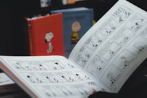 An open comic book displaying black and white comic strips. In the background, there are two hardcover books with colorful covers featuring characters from the comic. The image suggests a setting focused on reading or comic collection.