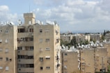A series of beige residential apartment buildings with multiple windows and satellite dishes on the rooftops. The scene is set against a backdrop of a partly cloudy sky. The buildings are surrounded by greenery, including trees and shrubs, and there are no visible people in the image.