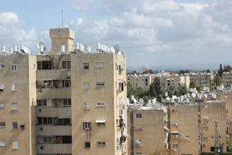 A series of beige residential apartment buildings with multiple windows and satellite dishes on the rooftops. The scene is set against a backdrop of a partly cloudy sky. The buildings are surrounded by greenery, including trees and shrubs, and there are no visible people in the image.