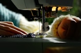 A close-up view of a sewing machine actively stitching fabric with visible hands guiding the material. The machine's needle and presser foot are in sharp focus as natural light brightens the scene.