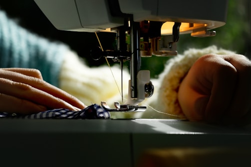A close-up view of a sewing machine actively stitching fabric with visible hands guiding the material. The machine's needle and presser foot are in sharp focus as natural light brightens the scene.