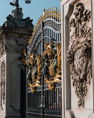 Ornate iron gates with intricate golden embellishments. The design includes detailed sculptures and decorative patterns. Stone structures with elaborate carvings flank the gates, depicting various symbols and figures.