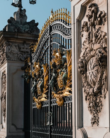 Ornate iron gates with intricate golden embellishments. The design includes detailed sculptures and decorative patterns. Stone structures with elaborate carvings flank the gates, depicting various symbols and figures.